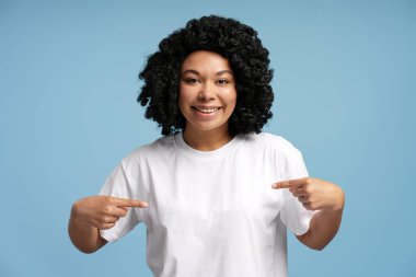 Kıvırcık saçlı, gülümseyen ve tişörtünü gösteren, mavi üzerine izole edilmiş kameraya bakan neşeli Afro-Amerikan kadının portresi. Yaşam tarzı, çeşitlilik ve duygu kavramı