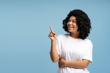 Kıvırcık saçlı, gülümseyen ve yukarıyı gösteren neşeli Afrikalı Amerikalı kadın, mavi arka planda soyutlanmış. Yaşam tarzı, çeşitlilik ve duygu kavramı