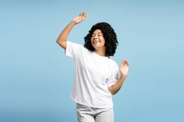 Beyaz bir tişört giyerken kıvırcık saçlı neşeli dans eden mutlu Afro-Amerikan kadının portresi. Eğlenceli konsept