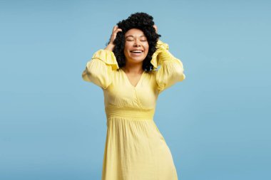 Sarı elbiseli mutlu Afro-Amerikan kadın portresi. Gözleri kapalı poz veriyor. Kıvırcık saçlarına dokunuyor. Saç stili konsepti