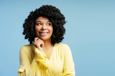 Gülümseyen Afro-Amerikalı genç bir kadının portresi, mavi arka planda tek başına duran bir kenara bakıyor. Uzayı kopyala Moda, güzellik, duygu konsepti