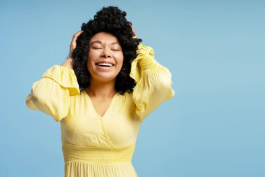 Gülümseyen güzel Afro-Amerikan kadın portresi. Gözleri kapalı, saçlarına dokunuyor. Mavi arka planda izole bir poz veriyor. Moda, güzellik, duygu konsepti