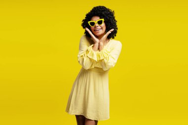 Sarı elbiseli, şık güneş gözlüklü, kıvırcık saçlı, yüzünün yanında el ele tutuşan güzel Afro-Amerikan kadın portresi. Moda, güzellik, duygu konsepti