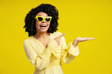 Kıvırcık saçlı, güneş gözlüğü takan ve sarı üzerine izole edilmiş işaret parmağıyla işaret eden neşeli Afro-Amerikan kadın portresi. Moda, güzellik, duygu konsepti