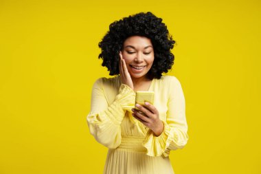 Sarı arka planda izole edilmiş, gülümseyen ve akıllı telefon tutan Afro-Amerikan kıvırcık saçlı genç kadının portresi. Yaşam tarzı, duygu, iletişim konsepti