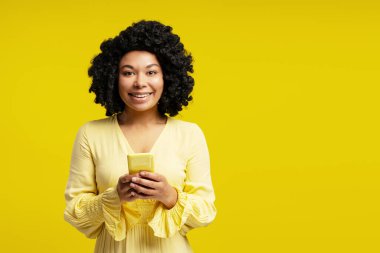 Sarı arka planda izole edilmiş akıllı telefondan mesaj atan mutlu kıvırcık kadın portresi, kameraya bakıyor, kopyalama alanı. Yaşam tarzı, duygu, iletişim konsepti