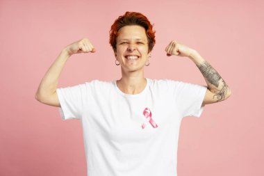 Pembe arka planda pazılarını gösteren pembe kurdeleli gülümseyen kızıl saçlı kadının portresi, meme kanseri farkındalığı konsepti.
