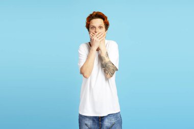 Şok olmuş genç bir adam, boyalı kızıl saçlı, ağzı ellerle kaplanmış, şaşırmış ya da mavi arka plandaki korkuyu ifade ediyor.