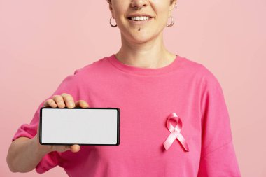 Göğüs kanseri farkındalığı için pembe kurdeleli pembe tişörtlü, beyaz ekranlı akıllı telefon gösteren bir kadın.