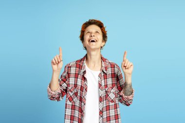 İşaret parmaklarıyla yukarıyı gösteren ve mavi arka planda heyecanla bakan neşeli genç bir kadının stüdyo portresi.