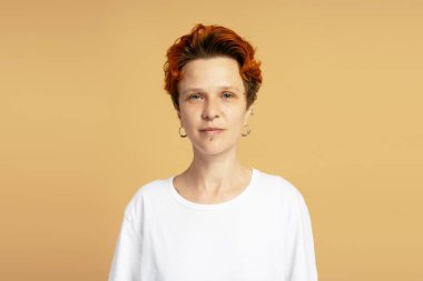 Çift cinsiyetli bir kişinin, bej ve turuncu arkaplan üzerine beyaz bir tişört giyerken özgüvenini gösteren bir stüdyo resmi.