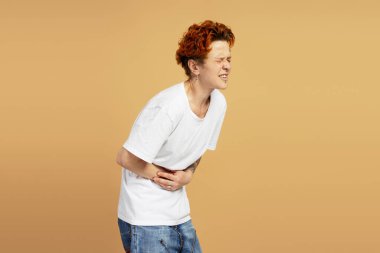 Piercing ve kızıl saçlı, karın ağrısı çeken, elleriyle karnını tutan ve bej arka planda rahatsız bir şekilde eğilen genç bir adam.