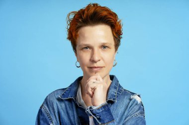 Boyalı turuncu saçlı ve piercingli, elleri çenesinde kameraya bakan kendinden emin genç bir kadının portresi.