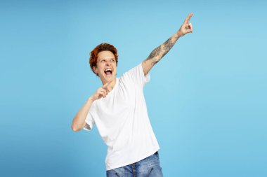 Turuncu saçlı, dövmeli, beyaz tişörtlü ve mavi arka planda gülüşen genç insanların stüdyo fotoğrafları.