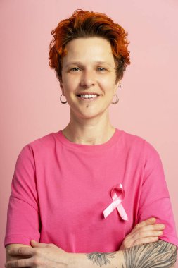 Pembe saçlı, pembe kurdeleli pembe tişörtlü ve göğüs kanseri farkındalığını destekleyen gülümseyen ikili olmayan bir kişinin portresi.