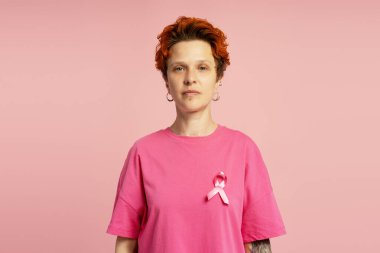 Kısa kızıl saçlı, pembe tişörtlü, göğüs kanserini destekleyen pembe kurdeleli ciddi bir yetişkin kadın pembe arka planda duruyor.
