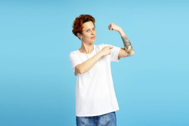 Kendine güveni olan bir gencin mavi arka planda dövmesi olan pazıları işaret ettiği bir stüdyo fotoğrafı.