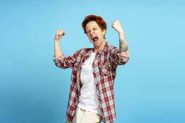 Kızıl saçlı tasarımcı sevinç ve heyecanı ifade ediyor, başarıyı sevinçli yumruklarla ve mavi arka planda neşeli bir ifadeyle kutluyor.