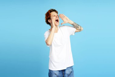 Dövmeli kızıl saçlı adam elleri ağzında bağırıyor, canlı mavi bir arka planda heyecan ve gürültüyü ifade ediyor.