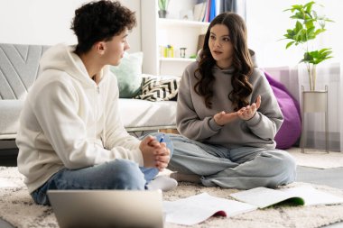 İki genç öğrenci yerde oturmuş ödevlerini tartışıyorlar. Yanında da dizüstü bilgisayar ve defterler var.