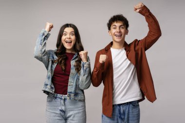 İki neşeli öğrenci akademik başarılarını yumruklarını kaldırarak ve ağızlarını açarak kutluyor. Gri bir arka planda neşe ve heyecan ifade ediyorlar.