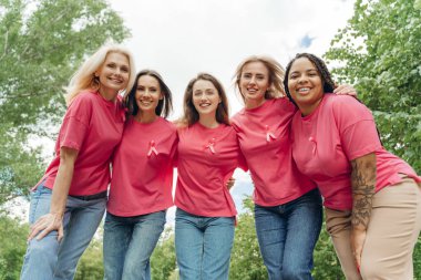 Farklı yaş ve etnik kökenlere sahip gülümseyen bir grup kadın açık havada kucaklaşıyor, pembe kurdeleli pembe tişörtler giyiyor ve meme kanseri farkındalığını arttırıyor.