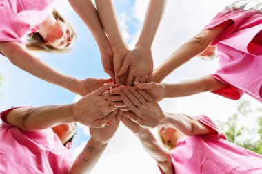 Pembe tişört giyen bir grup kadın ellerini bir daire şeklinde birleştirerek, arka planda açık bir gökyüzü bulunan göğüs kanseri için birlik, destek ve farkındalığı sembolize ediyor. Takım konsepti