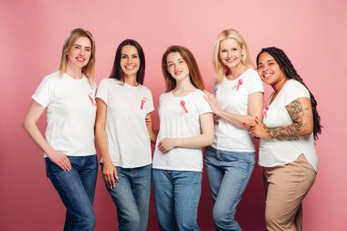 Çeşitli yaş ve etnik kökenlere sahip beş gülümseyen kadın, pembe kurdeleli beyaz tişörtlerle birleşerek, meme kanseri farkındalığı ve hastalığa karşı dayanışma gösteriyorlar.