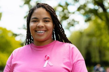 Pembe bir tişört giyen, göğüs kanseri farkındalığını sembolize eden pembe kurdeleli mutlu, Afro-Amerikan kadın parkta neşeyle gülümsüyor. Sağlık hizmeti kavramı