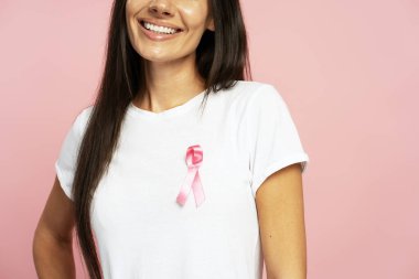 Beyaz tişörtüne pembe kurdele takmış gülümseyen bir kadının yakın çekimi göğüs kanseri farkındalığını sembolize ediyor ve umut ve dayanışmayı ifade ediyor.
