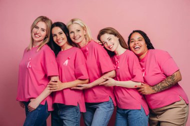 Pembe kurdeleli pembe tişörtler giyen beş tane gülümseyen çok ırklı kadın göğüs kanseri farkındalığını desteklemek için birbirlerine sarılıyorlar, pembe bir arka planda birlik ve umut gösteriyorlar.