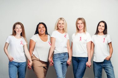 Farklı yaşlardan beş gülümseyen kadın el ele tutuşuyor ve pembe kurdeleli beyaz tişörtler giyiyor, stüdyo ortamında meme kanseri farkındalığını sembolize ediyor. Sağlık hizmeti kavramı