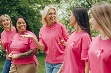 Pembe kurdeleli pembe tişörtler giyen beş farklı kadın parkta birlikte yürüyor, sohbet ediyor ve el kol hareketi yapıyor, meme kanseri farkındalığını destekliyorlar.