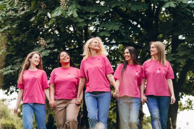 Göğüs kanseri farkındalığını sembolize eden pembe kurdeleli pembe tişörtler giyen beş farklı kadın, el ele tutuşup parkta birlikte yürüyorlar, birlik ve destek gösteriyorlar. Sağlık hizmeti kavramı