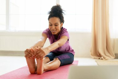 Mutlu atletik kadın evde yoga yapıyor, dizüstü bilgisayarda özel ders izliyor ve yoga minderinde bacaklarını açıyor.