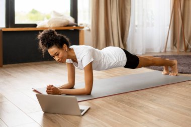 Spor kadını dizüstü bilgisayarda online fitness antrenmanı izlerken tahta egzersizi yapıyor, evde sağlıklı bir yaşam sürüyor.
