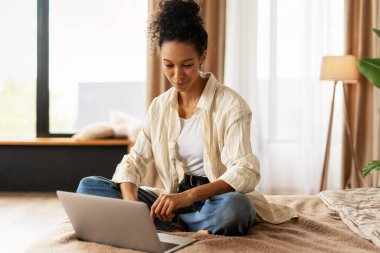 Yatakta rahat bir şekilde otururken dizüstü bilgisayar kullanan gülümseyen kadın, uzaktan çalışmanın ve ev ofisinin keyfini çıkarıyor.