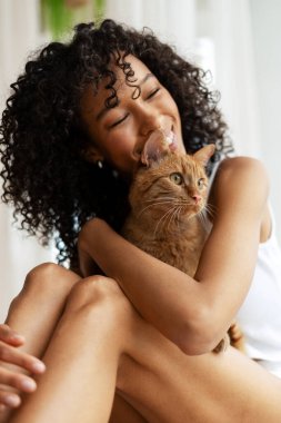 Mutlu genç bir kadın, sevimli kızıl kedisine sarılıp evinde rahat bir şekilde otururken sıcak bir ortamda neşe ve şefkat yayıyor.