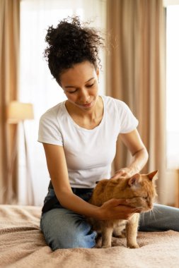 Gülümseyen bir kadın evinde rahat ve huzurlu bir anın tadını çıkarırken bir yatakta oturmuş kızıl bir kediyi nazikçe okşuyor.