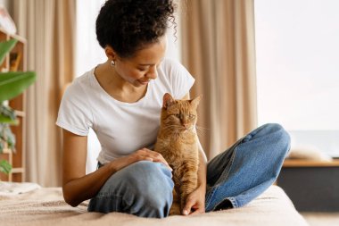 Mutlu kadın sahip bacak bacak üstüne atmış, kızıl kedisini okşayıp kucaklıyor, evde birlikte vakit geçiriyorlar.