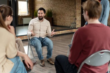 Rehabilitasyon merkezinde grup terapisi sırasında hastalarla konuşurken el kol hareketi yapan sakallı bir terapist.