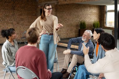 Gülümseyen koç, tekerlekli sandalyedeki yaşlı bir adam da dahil olmak üzere çeşitli insanlarla grup toplantısına liderlik ederken parmağını işaret ediyor.