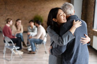 Genç kadın yaşlı babasını kucaklıyor, grup terapisi sırasında ona sevgi ve destek sağlıyor.