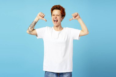 Beyaz tişörtlü, poz veren ve çığlık atan, baş parmaklarını gösteren, mavi üzerine izole genç, duygusal tuhaf insan. Bireysellik, çeşitlilik ve kendini ifade etme kavramı