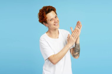 Heyecanlı kadın el çırpıyor ve kameraya mutlu bir şekilde gülümsüyor. Mavi arka planda beyaz tişörtle dikiliyor. Bireysellik, çeşitlilik ve kendini ifade etme kavramı