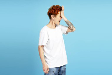 Depresyonda olan, piercing giyen beyaz tişörtlü, alnına el ele tutuşmuş, mavi arka planda izole edilmiş bir kadın. Bireysellik, çeşitlilik ve kendini ifade etme kavramı