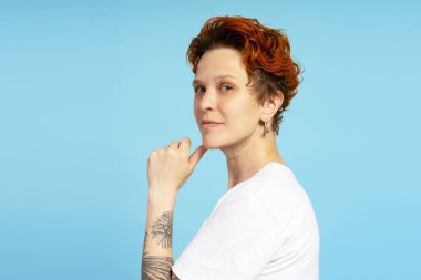 Kol dövmeli, beyaz tişörtlü, düşüncelere dalmış, mavi arka planda soyutlanmış kızıl saçlı bir kadın. Bireysellik, çeşitlilik ve kendini ifade etme kavramı