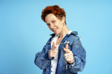 Kısa kızıl saçlı ve piercingli gülümseyen genç bir kadının portresi, kamerayı işaret ediyor, mavi üzerine izole edilmiş. Bireysellik, çeşitlilik ve kendini ifade etme kavramı