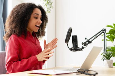 Profesyonel mikrofon ve dizüstü bilgisayar kullanarak ses içeriğini kaydeden coşkulu genç kadın, evden çalışıyor.