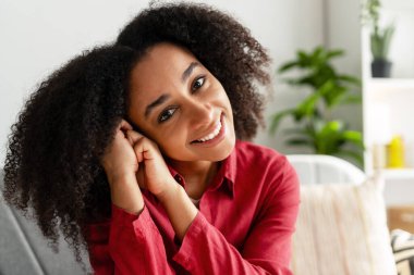 Kıvırcık saçlı neşeli bir kadın. Parlak bir oturma odasında huzurlu bir anın tadını çıkarıyor. Etrafı bitkilerle ve rahat dekorla çevrili.